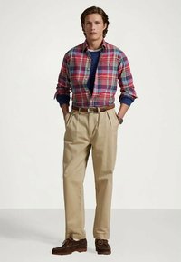 Raštuota marškiniai raudonos, mėlynos ir baltos spalvos, dėvimi virš tamsiai mėlyno megztinio. Komplektą sudaro smėlio spalvos kelnės, ruda diržas ir tamsiai rudi bateliai loaferiai.