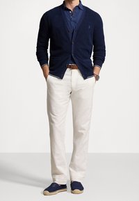 Marinblå stickad kofta över en marinblå polo, krämfärgade byxor och marinblå espadriller med juteaccent. Knappdetaljerna på koftan är synliga.