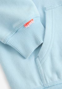 Maglione azzurro chiaro in tessuto con polsini a coste. Presenta un'etichetta rossa con la scritta "by superdry." Texture morbida, design semplice e stile a maglione.