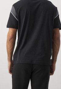 Svart T-shirt med grå axel detaljer och vit piping. Korta ärmar, rund halsringning, tillverkad av bomull. Bärs med svarta byxor.