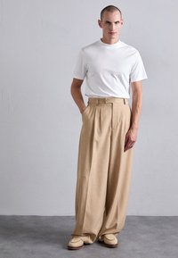 Mann som står mot en grå vegg iført en hvit kortermet skjorte, beige vide bukser og kremfargede vevde loafers, med hendene i lommene.
