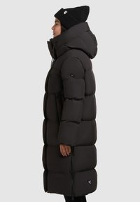 Sort dunjakke med hætte, der har et firkantet design, quiltet tekstur og en lynlås på siden. Bæres med en sort strikbeanie.