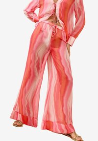 Zwei-teiliger Anzug aus leichtem, transparenter Stoff. Das Oberteil ist pink mit gewellten Mustern und Schlaufenbändern; die Hose ist ausgestellt und passt mit einem ähnlichen Design dazu.