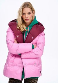 Różowa kurtka puchowa z bordowym nałożeniem i dużym kapturem. Posiada zapięcie na zatrzaski oraz pikowany design. Nosi się ją na zielonej bluzie z kapturem.