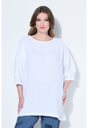 Femme aux cheveux bruns mi-longs portant un haut blanc texturé surdimensionné et un pantalon bleu à imprimé floral foncé, debout devant un fond blanc.