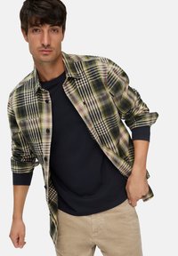 Man in een groen en zwart geruite shirt over een zwart langemouwen shirt, met beige broek, poseert tegen een effen achtergrond.