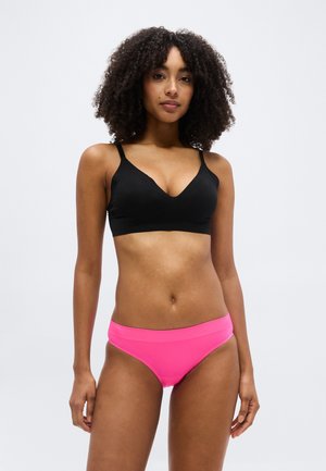 Femme aux cheveux bouclés portant un soutien-gorge noir et une culotte rose vif, debout devant un fond gris clair uni.