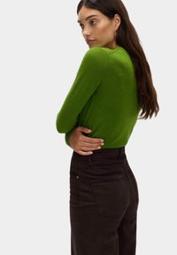 Kvinna med långt mörkt hår, klädd i grön långärmad topp och höga svarta byxor, som ser över axeln mot en enhetlig bakgrund.