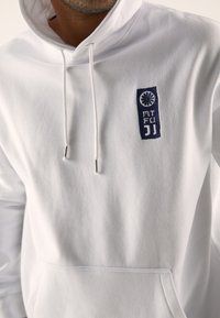 Felpa bianca realizzata in morbido misto cotone, con tasca frontale, cordini e un logo ricamato blu navy con un emblema decorativo.