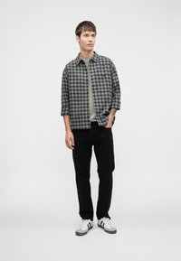 Καρό πουκάμισο σε μαύρο και γκρι, με κουμπιά, κοντό μανίκι, πάνω από ένα ανοιχτό πράσινο t-shirt. Μαύρο παντελόνι και γκρι-λευκά αθλητικά παπούτσια.