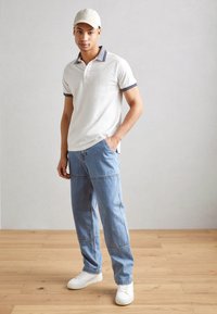 Cămașă polo albă cu guler în dungi bleumarin, pantaloni din denim deschis și adidași albi. Modelul stă pe un podea din lemn, în fața unui fundal gri deschis.
