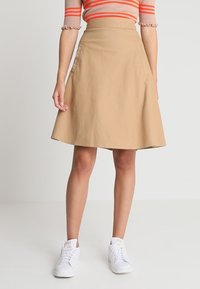 Una falda A-line color caqui hecha de sarga de algodón, con bolsillos laterales y una textura suave, combinada con zapatillas blancas.