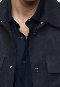 Námořnicky modrá semišová bunda s patentními knoflíky a dvěma předními kapsami, vrchní vrstva přes tmavě modrou košili s límečkem na knoflíky a kontrastní texturou.