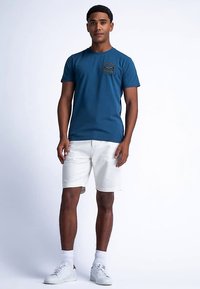 Modré bavlněné tričko s malým grafickým motivem na přední straně, doplněné o bílé šortky a bílé tenisky. Model stojí před jednobarevným pozadím.