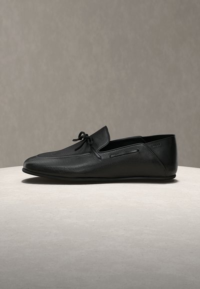 Mocassin en cuir noir avec couture moc toe et détail nœud sur une surface claire et un fond neutre.
