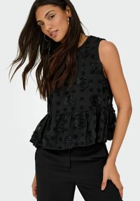 Femme portant un haut noir sans manches à motifs floraux texturés avec un ourlet à volants et un pantalon noir, posant avec les cheveux détachés et des boucles d'oreilles créoles.