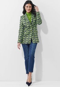 Groene en marineblauwe blazer met bloemendessin in een getailleerde pasvorm, gecombineerd met een groene coltrui en blauwe jeans, enkellang, met puntige hakken.