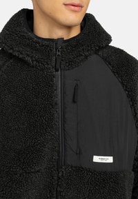 Uomo che indossa una giacca in pile nera con colletto alto e tasca sul petto con zip etichettata "ELEMENT CO. SINCE 1992"