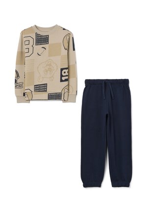 Sweatshirt beige à motifs avec un pantalon de jogging bleu marine. Le sweatshirt présente des graphiques et du texte liés au sport ; le pantalon a une taille élastique et des poignets élastiques.