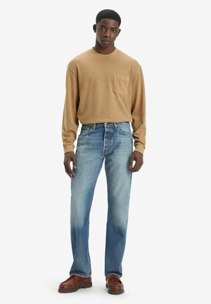Beige overhemd met lange mouwen en een borstzak, lichtblauwe jeans, bruine schoenen. Rechte denim met subtiele vervaging; casual pasvorm.