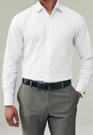 Homme portant une chemise blanche à manches longues rentrée dans un pantalon gris avec une ceinture noire, main dans la poche, tête non visible.