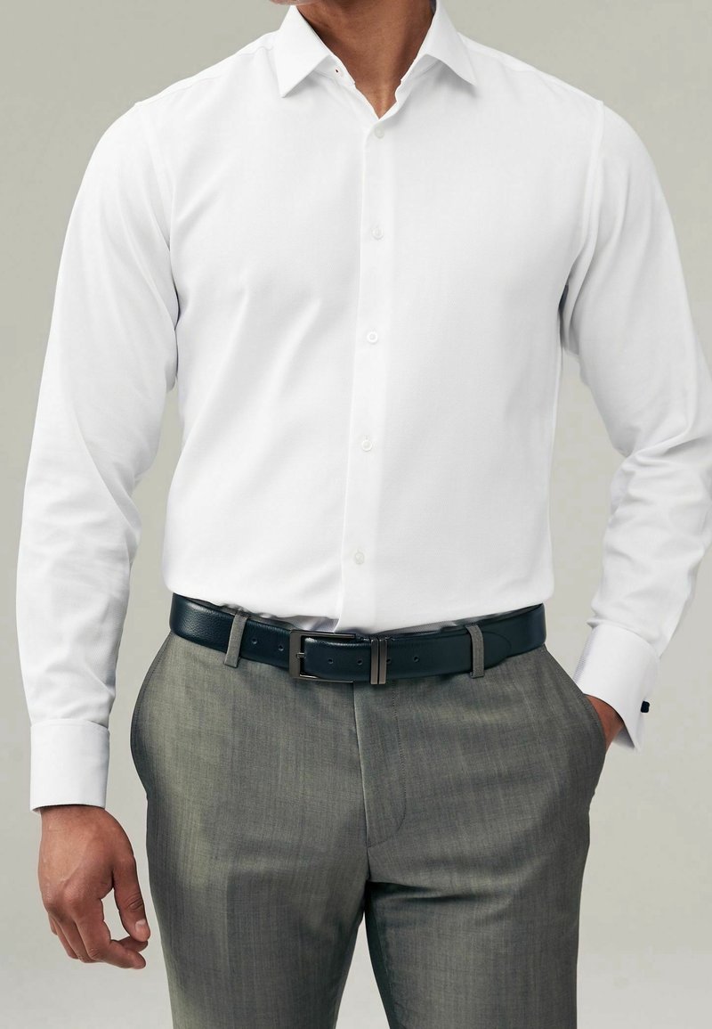 Homme portant une chemise blanche à manches longues rentrée dans un pantalon gris avec une ceinture noire, main dans la poche, tête non visible.