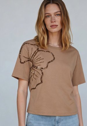 Frau mit schulterlangem blonden Haar, die ein braunes T-Shirt mit einem großen Pailletten-Schmetterlingsdesign auf der linken Seite und hellblaue Jeans trägt.