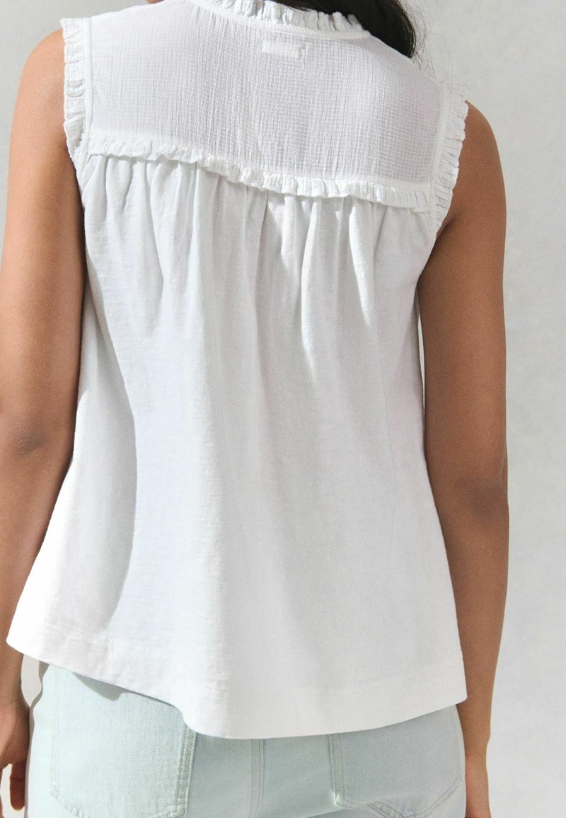 Blouse blanche sans manches avec un empiècement volanté aux épaules, portée par une personne vue de dos sur un fond uni.
