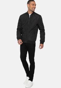 Zwarte gewatteerde bomberjack met een rits aan de voorkant en zijzakken, gecombineerd met zwarte slim-fit broek en witte sneakers.