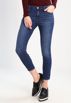 Frau trägt dunkelblaue Skinny-Jeans, schwarze Plateau-Schnürschuhe und ein dunkelrotes Langarmtop, eine Hand in der Tasche.