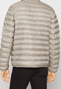 Giubbotto imbottito grigio chiaro con design a trapuntatura orizzontale, caratterizzato da una superficie liscia e lucida. Chiusura frontale con zip e polsini elastici.