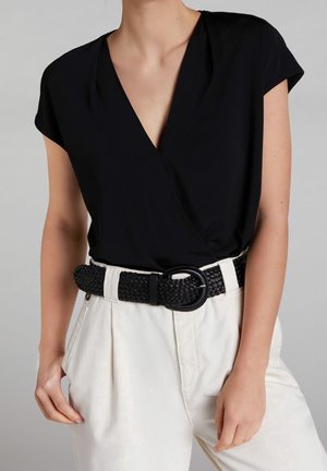 Femme portant un haut noir à manches courtes et col en V, rentré dans un pantalon blanc taille haute avec une ceinture noire tissée.