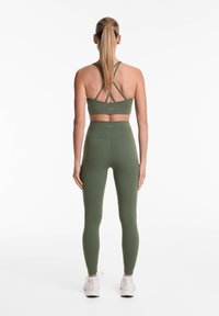 Zelena športna modrček z x-ovimi trakovi, vparen z visokimi zelenimi legginsami. Gladka tekstura, minimalistična zasnova, diskretno znamčenje na pasu.