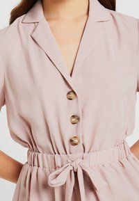 Vestito rosa chiaro con colletto, tre bottoni e vita segnate da una cintura. Presenta un tessuto testurizzato e maniche corte.