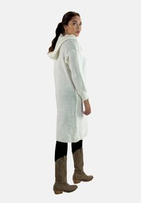 Vestido blanco de punto con capucha y textura acanalada, que presenta mangas largas y un corte hasta la rodilla, combinado con leggings negros y botas de vaquero color miel.