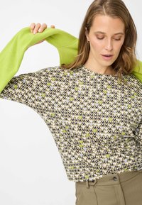 Top met lange mouwen en een boxy pasvorm, met een golvend patroon in beige, bruin en groen, gecombineerd met een felgroene trui die over de schouders is gedrapeerd.