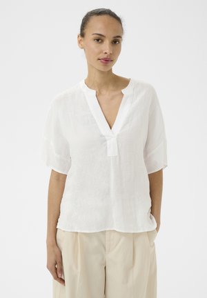 Femme aux cheveux attachés portant un chemisier blanc ample à col en V et un pantalon beige, debout devant un fond blanc uni.