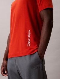 T-shirt de algodão vermelho com mangas curtas e logótipo branco "Calvin Klein" na lateral, combinado com calças cinzas texturizadas com uma etiqueta da marca.