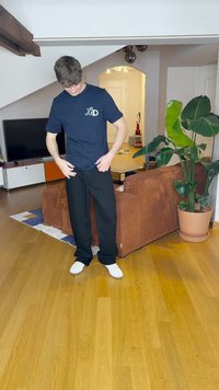 Μαύρο παντελόνι και ναυτικό μπλουζάκι με λευκό λογότυπο, φορεμένο με λευκά παπούτσια. Εσωτερικός χώρος με ξύλινο πάτωμα και ένα πράσινο φυτό.