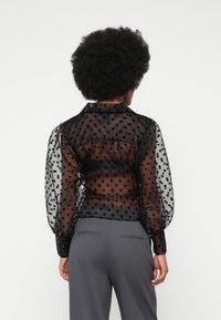 Blouse noire transparente avec un motif à pois, manches bouffantes et col. Stylisée avec un pantalon gris, mettant en valeur une silhouette ajustée.