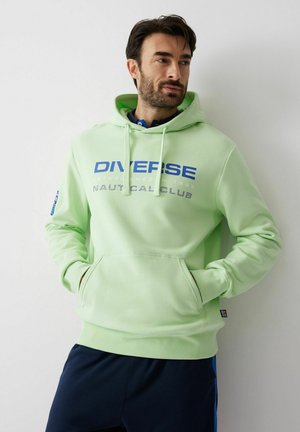 Mężczyzna w jasnozielonej bluzie z kapturem z napisem "DIVERSE NAUTICAL CLUB", ręce w przedniej kieszeni, stoi przy białej ścianie.