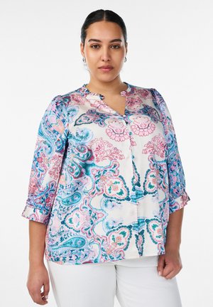 Žena oblečená v bielu blúzku s modrými a ružovými kvetinovými vzormi paisley a bielymi nohavicami, stojaca pred jednofarebným pozadím.