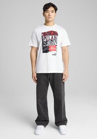Camiseta blanca de manga corta con un diseño gráfico en negro y rojo que incluye el texto "Forza Milan", combinada con unos vaqueros gris oscuro y zapatillas blancas.
