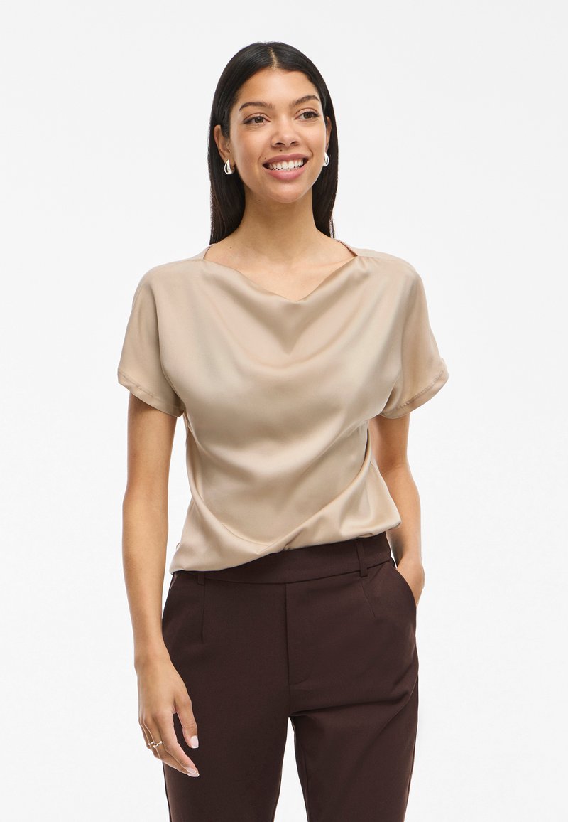 Donna sorridente con capelli lunghi e scuri indossa una camicia beige in raso a maniche corte e pantaloni marrone scuro, in piedi con una mano in tasca.