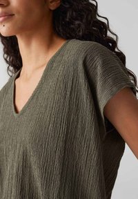 Olive green top with a textured, crinkled fabric, featuring a V-neck and short sleeves, designed for a relaxed fit.