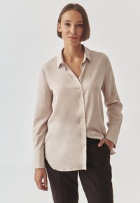 Light beige button-up shirt made of smooth fabric, featuring a pointed collar, long sleeves, and a curved hemline, paired with dark pants.