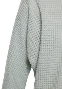 Nahaufnahme von hellgrauem Waffle-Knit-Stoff auf einem Langarmkleidungsstück, das das detaillierte Texturmuster und die Naht unter dem Arm zeigt.
