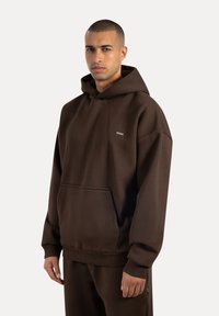 Man som bär en brun oversized hoodie med en framficka och matchande bruna byxor mot en enkel ljus bakgrund.