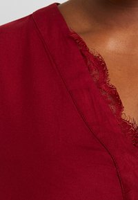 Indumento bordeaux con scollatura a V, caratterizzato da un delicato bordo in pizzo lungo il margine e una texture morbida e liscia.