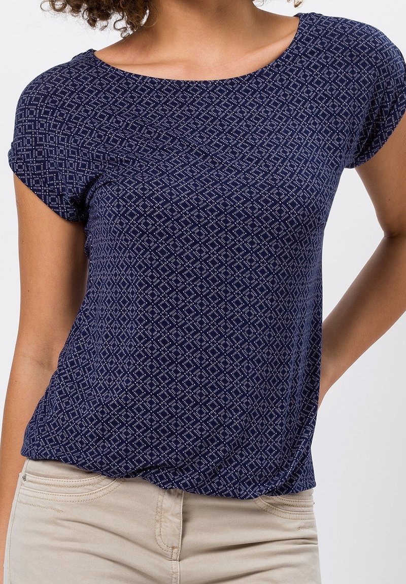 Kvinna i en marinblå kortärmad topp med ett litet vitt diamantsmönster och ljusbeiga byxor, med handen vilande på höfterna.